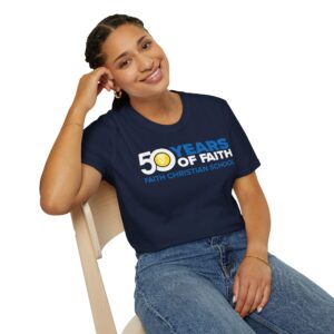 A smiling woman leaning on her hand, wearing a blue t-shirt with "50 YEARS OF FAITH, FAITH CHRISTIAN SCHOOL" printed on it, sitting on a wooden chair against a white background.