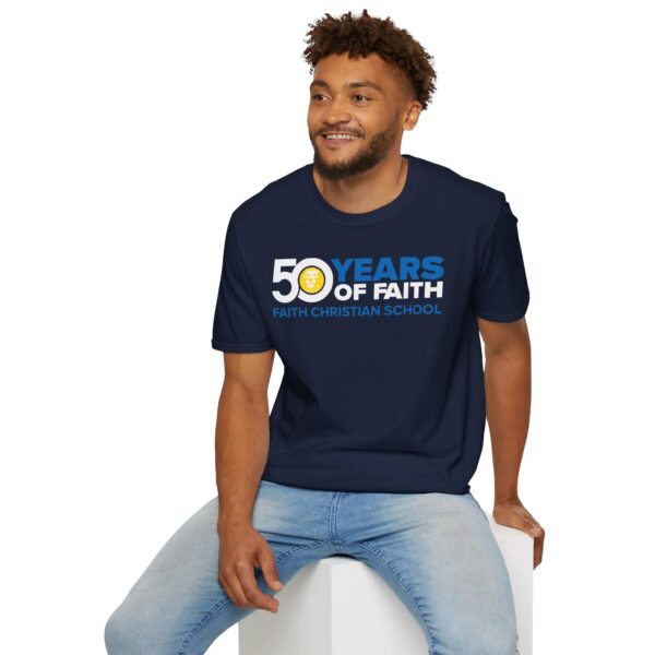 A man sitting on a white block, smiling slightly, and wearing a dark blue t-shirt with the text "50 YEARS OF FAITH - FAITH CHRISTIAN SCHOOL" printed on the front, paired with light blue jeans.